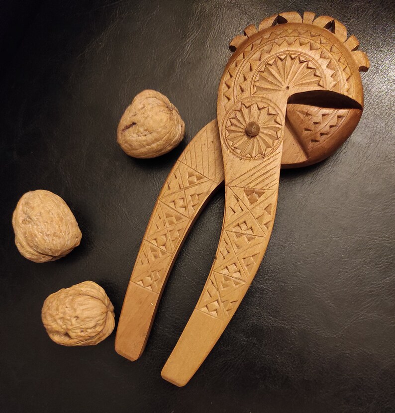 Op de afbeelding: Een houten notenkraker met ingewikkelde houtsnijwerk in de vorm van een vogel. De notenkraker is open en er liggen drie walnoten in de buurt.