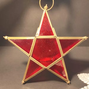 May include: A five-pointed star-shaped lantern constructed with red textured glass panels framed in brass-toned metal. The lantern features a circular metal ring at the top for hanging and pointed metal tips at each of the five star points.