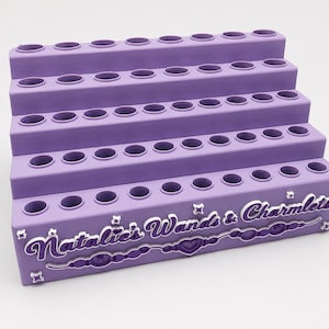May include: A lavender tiered display stand with five levels, each featuring a row of circular holes for storage. The front panel displays the white text "Natalie's Wands & Charmlets" above a decorative line of beads and a central heart shape.
