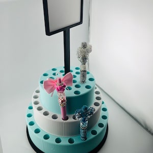 Op de afbeelding: Een penhouder met drie niveaus in blauw-, wit- en zwarttinten, met een zwart bord. De houder bevat decoratieve pennen met strikken en kristallen versieringen. De penhouder is ongeveer 20 cm hoog.