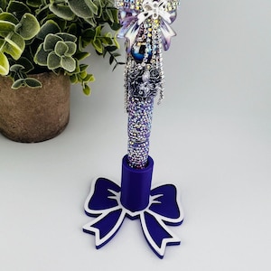 May include: A decorative pen with a purple and silver design. The pen features a jeweled cross and bow, with a purple base and a bow-shaped stand. The pen is covered in small, sparkling crystals.