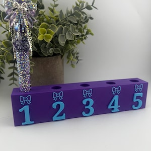 Puede incluir: Un portalápices morado con cinco agujeros, cada uno marcado con un número azul claro. El soporte tiene un diseño de lazo encima de cada número. Un bolígrafo enjoyado con un diseño morado y plateado está en el primer agujero.