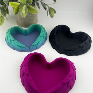 May include: Three heart-shaped trinket dishes with wing-like accents. One is a gradient of teal to blue, another is solid black, and the third is a vibrant magenta. These decorative dishes are ideal for holding small items.