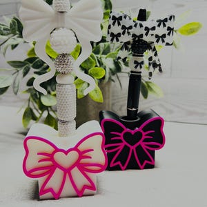 May include: Two pens with decorative toppers and matching pen holders. One pen is white with a white bow topper and a white holder featuring a hot pink heart and bow outline. The other is black with a black and white bow topper and a black holder.