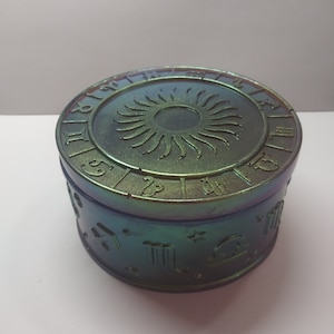 May include: A round, iridescent green and blue trinket box with a zodiac wheel design on the lid and zodiac symbols around the sides.