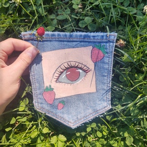 May include: A denim pocket with a painted eye and strawberries. The eye is green with a red iris and eyelashes. The strawberries are red with green leaves.