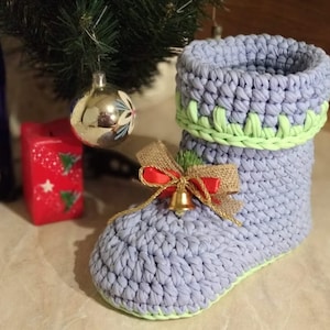May include: A crocheted boot-shaped container in light purple with a green trim. The boot has a gold bell and a red and brown bow attached to it.