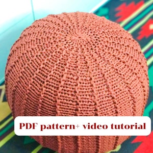 May include: A large, round, knitted ottoman in a terracotta color. The ottoman is sitting on a colorful rug with a geometric pattern. The text "PDF pattern + video tutorial" is displayed on a white rectangle in front of the ottoman.