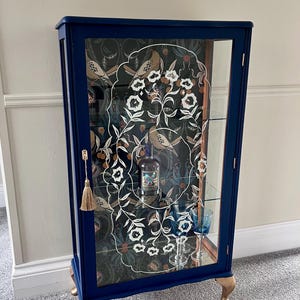 May include: A navy blue display cabinet with glass doors and shelves. The cabinet has gold-coloured feet and a decorative tassel. The interior features a patterned backing with birds and floral designs. A bottle and glasses are inside.