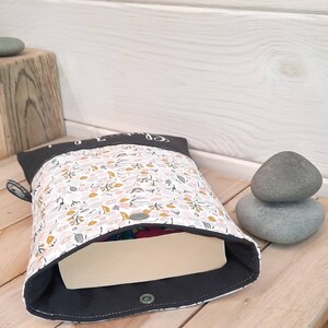 May include: A fabric book sleeve with a floral pattern in pink, yellow, and green on a white background. The sleeve has a dark gray border and a snap closure. A book is partially visible inside.
