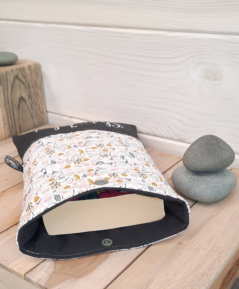 May include: A fabric book sleeve with a floral pattern in pink, yellow, and green on a white background. The sleeve has a dark gray border and a snap closure. A book is partially visible inside.