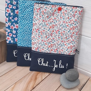 May include: Three fabric book sleeves with floral and leaf patterns. Each sleeve has a navy blue bottom panel with white script. The text on the right sleeve reads "Chut...Je lis!" A small red pom-pom adorns the top of one sleeve.