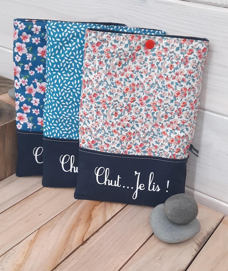 May include: Three fabric book sleeves with floral and leaf patterns. Each sleeve has a navy blue bottom panel with white script. The text on the right sleeve reads "Chut...Je lis!" A small red pom-pom adorns the top of one sleeve.