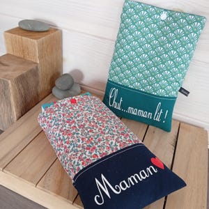 May include: Two fabric book sleeves. One features a floral pattern with red flowers and a navy blue bottom with the word "Maman" and a red heart. The other has a green and white fan design with the text "Chut...maman lit!"