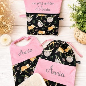 Peut inclure: Un ensemble composé d'une pochette zippée, d'un bavoir et d'un sac à cordon, tous en tissu noir à motif de paresseux et feuilles tropicales, avec des accents roses et le prénom Auria brodé en gris sur les parties roses.