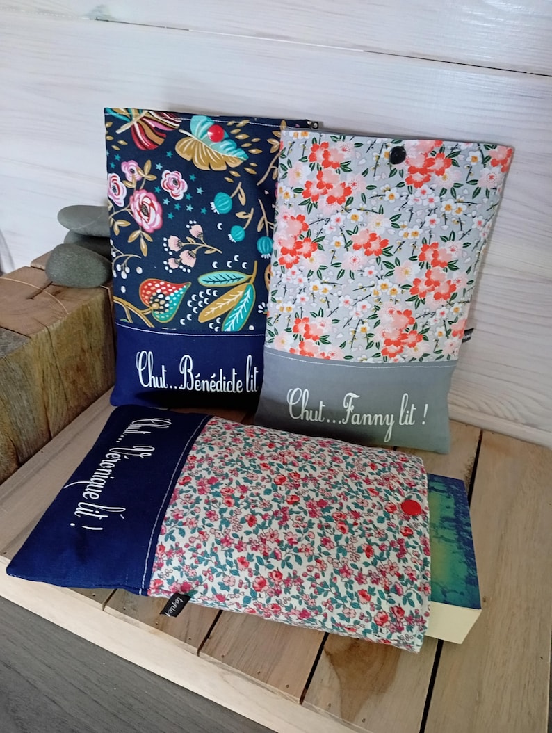 May include: Three fabric book sleeves with floral patterns and text. One sleeve is navy blue with a floral design and the text "Chut... B&eacute;n&eacute;dicte lit!". Another is gray with orange flowers and the text "Chut... Fanny lit!". The third is floral with a red button.