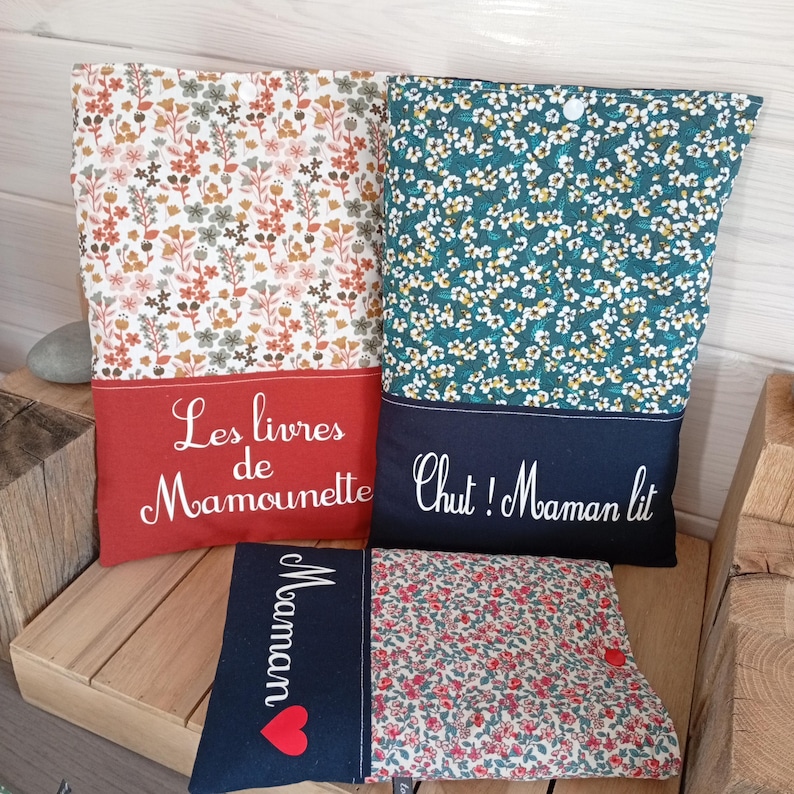 May include: Several fabric book covers with floral patterns and text. One cover has a red base with the text "Les livres de Mamounette". Another has a teal base with the text "Chut! Maman lit". A smaller cover has the word "Maman" and a heart.