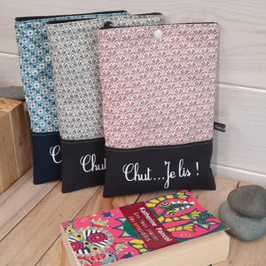 May include: Three fabric book covers in blue, gray, and pink patterns, each with a black bottom panel. The pink cover has the text "Chut...Je lis!" in white script. A book with a colorful cover is in front.
