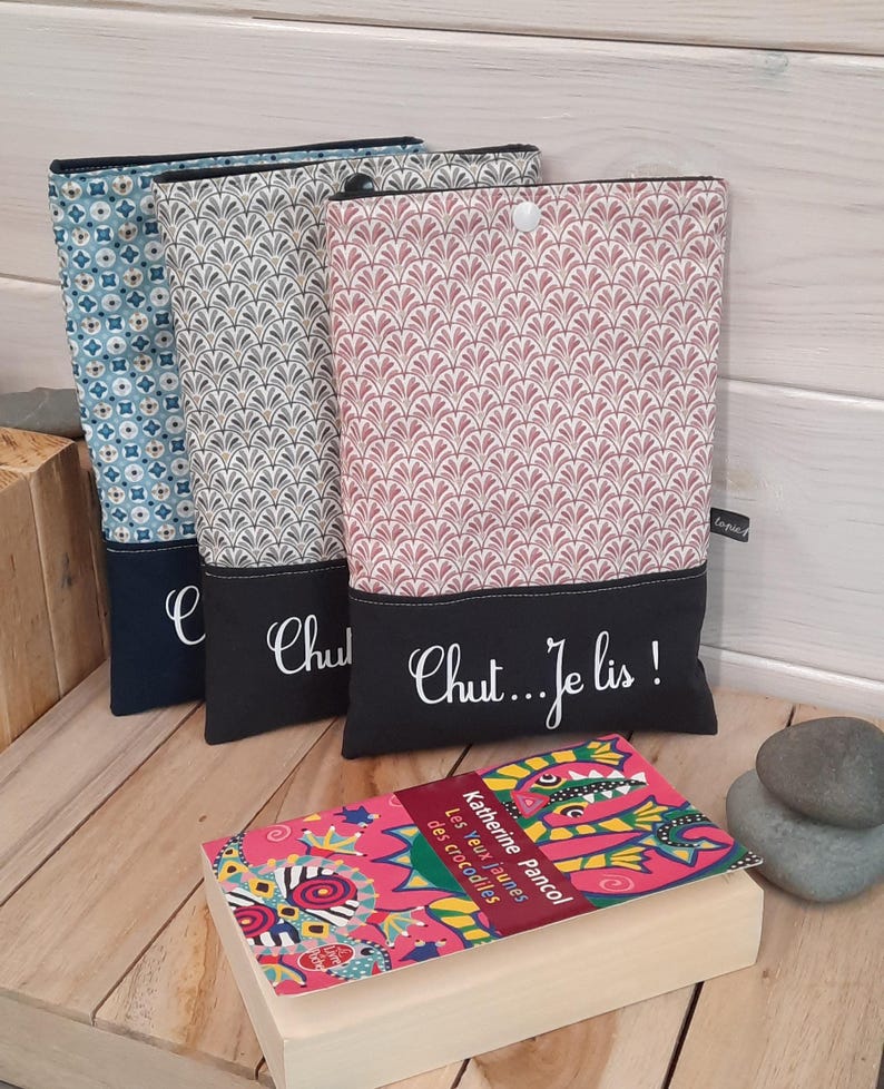 May include: Three fabric book covers in blue, gray, and pink patterns, each with a black bottom panel. The pink cover has the text "Chut...Je lis!" in white script. A book with a colorful cover is in front.