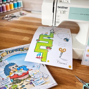 Peut inclure: Une machine à coudre Brother blanche avec un accent bleu clair coud une page illustrée colorée. Des bobines de fil et des ciseaux sont également sur la surface en bois. L'image présente un livre sur le thème de la couture.