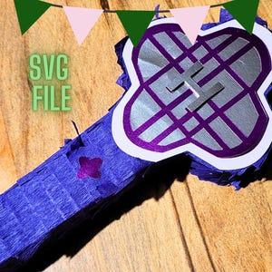 Puede incluir: Una piñata hecha a mano con una forma única, con un diseño morado y plateado con un borde blanco. La piñata está hecha de papel crepé y tiene el texto "SVG FILE" en verde. Se ven banderines decorativos en el fondo.