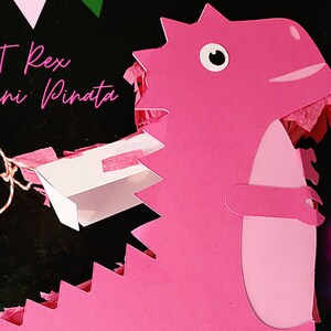 Könnte beinhalten: Rosa T-Rex Mini-Piñata mit einem weißen Papiereinsatz und einer Schnur zum Aufhängen. Die Piñata ist aus Papier und hat eine strukturierte, fransige Optik.  SVG-DATEI.