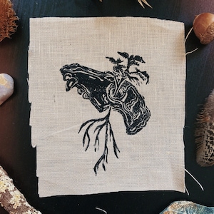 May include: A beige fabric square featuring a black linocut print of a tree trunk with roots and leaves. The image is surrounded by natural items such as a feather, acorn, and dried plants, giving a rustic, earthy feel.