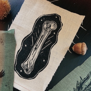Peut inclure: Une impression linogravée en noir et blanc d'un os sur un morceau de tissu de lin naturel. Le tissu a des bords bruts et est entouré d'autres morceaux de tissu avec des imprimés botaniques, un gland et une gousse de graine épineuse.
