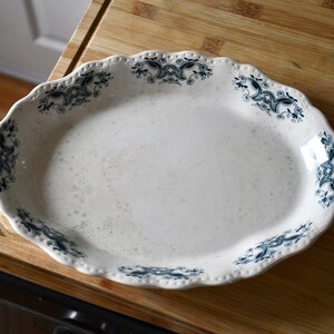 May include: Oval, white ceramic serving platter with a scalloped edge. The platter features a blue floral design around the rim. The platter is on a wooden surface.