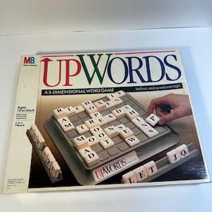 May include: A vintage Upwords word game. The box features the game title in red and green, with the tagline "Spell out, stack up and score high!". The game board is gray, with letter tiles and a hand placing a tile. Ages 10 to Adult, 2 to 4 Players.