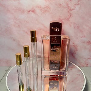 May include: A perfume bottle and three small spray bottles with gold caps. The perfume bottle is rectangular with a gold and brown cap. The bottles are on a mirrored surface with a pink marble background.