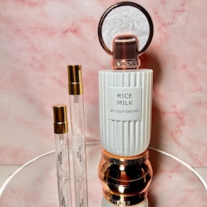 May include: A white perfume bottle with a ribbed design, a copper-colored cap, and the text "RICE MILK BY GULF ORCHID." Two small glass vials with gold-colored caps are next to the bottle. The items are on a mirrored surface against a pink marble background.