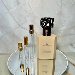 May include: A beige perfume bottle with a black decorative top and the text "SWISS ARABIAN VANILLA 01 EXTRAIT DE PARFUM." Three small, clear glass vials with gold caps are next to the bottle. The items are on a mirrored tray.