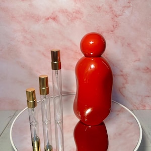 May include: A collection of perfume bottles and atomizers. The image features three clear glass atomizers with gold-colored caps and a large, red, rounded perfume bottle. The items are arranged on a mirrored surface.