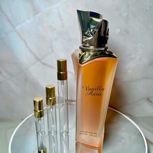 May include: A perfume bottle and several small spray bottles are displayed on a mirrored surface. The perfume bottle is a gradient of orange and peach with a gold cap and the words "Vanilla Aura" printed on the front. The small bottles have gold caps.