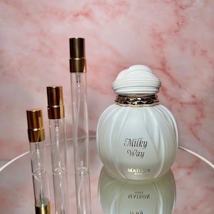 May include: A white perfume bottle with the text "Milky Way" and three clear glass spray bottles with gold caps. The perfume bottle has a unique, textured design and sits on a reflective surface. The background is a pink marble pattern.