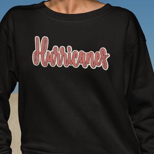 May include: Black sweatshirt featuring the word "Hurricanes" in red with a white outline. The text has a textured, embroidered look. The background is a gradient of blue.