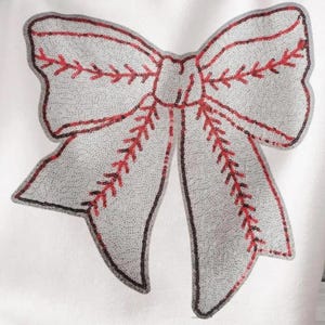 Puede incluir: Un lazo blanco con costuras rojas, que se asemeja a una pelota de béisbol, está hecho de lentejuelas. El lazo es un adorno decorativo para la ropa o los accesorios.