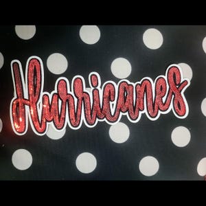 May include: A sticker with the word "Hurricanes" in red sequined cursive lettering, outlined in white. The background is black with white polka dots. A decorative item.
