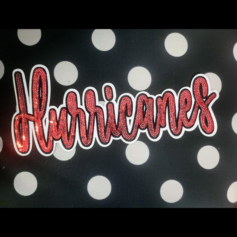 May include: A sticker with the word "Hurricanes" in red sequined cursive lettering, outlined in white. The background is black with white polka dots. A decorative item.
