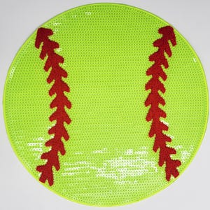 Pode incluir: Uma bola de softball verde e vermelha com costuras brancas. A bola de softball é feita de lantejoulas e tem costuras vermelhas felpudas.
