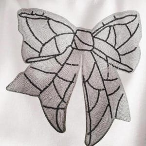 May include: A white fabric with a large, black and white, beaded bow applique. The bow is made of small, round beads that are sewn together to create a detailed design.