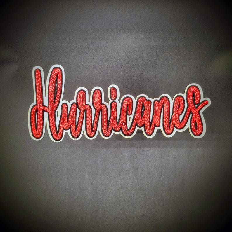 May include: A sticker featuring the word "Hurricanes" in red script. The letters have a glittery texture and are outlined in black and white. The sticker is set against a dark grey backdrop.