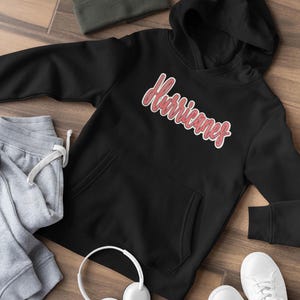 May include: Black hoodie featuring the word "Hurricanes" in red script. The hoodie has a front pocket and a hood. Grey joggers, a beanie, white headphones, and white trainers are also visible.
