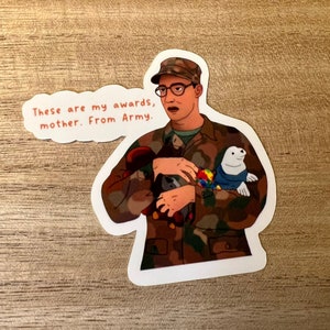 Puede incluir: Una pegatina de dibujos animados de un hombre con uniforme militar que sostiene animales de peluche. El texto de la pegatina dice "Estos son mis premios, madre. Del ejército."