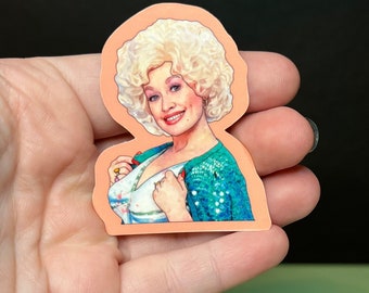 May include: A sticker featuring a portrait of a woman with blonde curly hair, wearing a green sweater and a floral top. The sticker has a peach-colored border and is held in a hand.