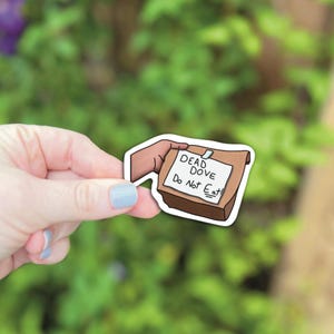 Puede incluir: Una pegatina que representa una bolsa de papel marrón con una nota que dice "DEAD DOVE Do Not Eat!". La pegatina está sujeta por una mano con una uña pintada de azul claro, sobre un fondo de hojas verdes.