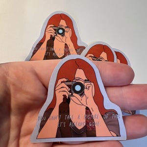 Könnte beinhalten: Ein Aufkleber mit einer Frau mit roten Haaren, die eine Kamera hält. Der Aufkleber trägt den Text "You can't take a picture of this, it's already gone."