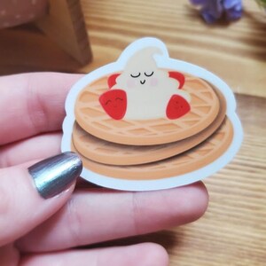 May include: A sticker of a stack of two waffles with a smiling whipped cream topping and two red strawberries. The waffles are brown and the whipped cream is white.