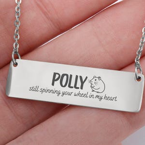 May include: A silver-colored bar necklace with a chain. The bar is engraved with the name "POLLY", a small hamster graphic, and the phrase "still spinning your wheel in my heart".